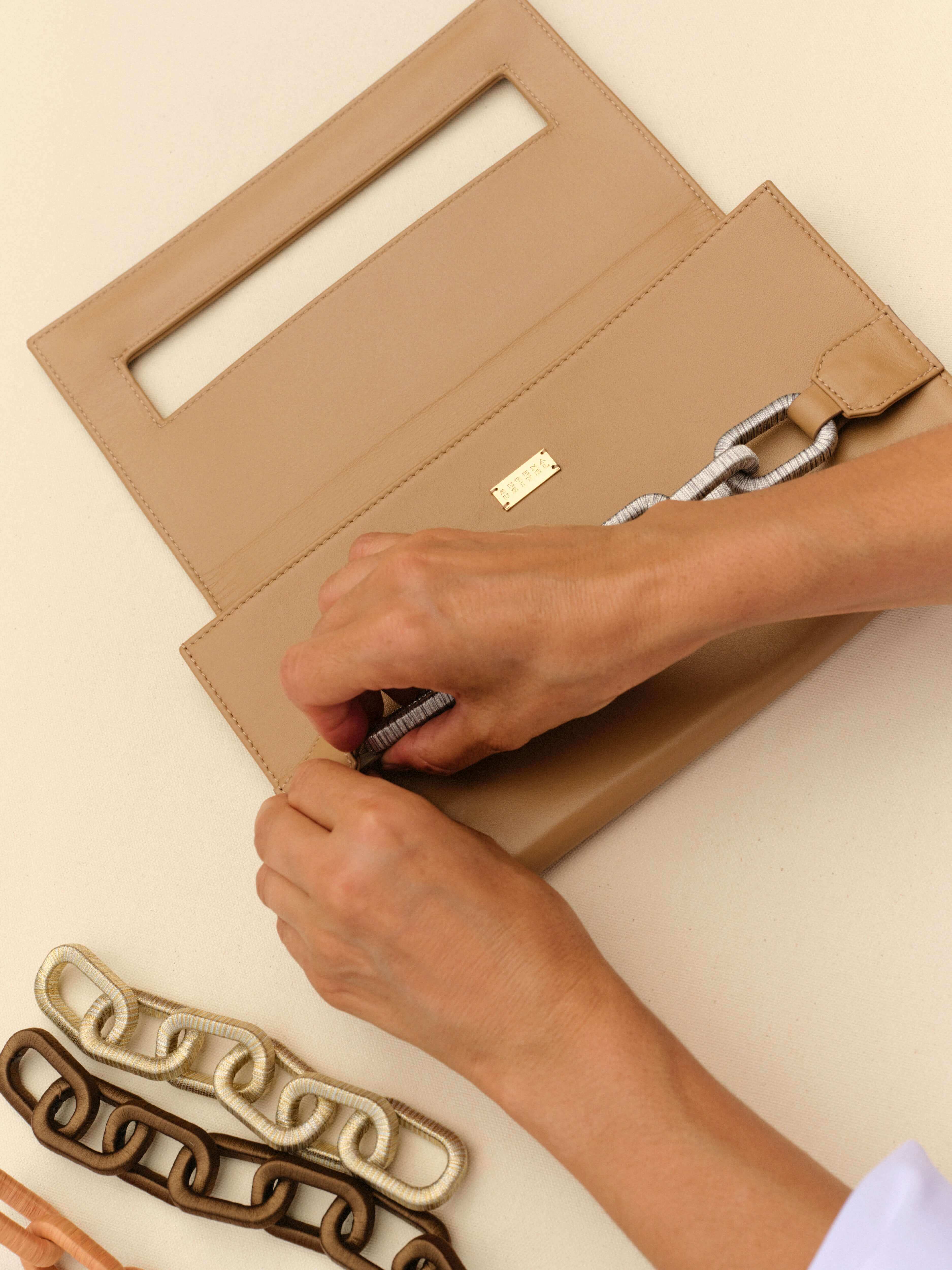 Person's hands adjusting a clasp on a beige purse with decorative chains laid beside it, showcasing style and elegance.