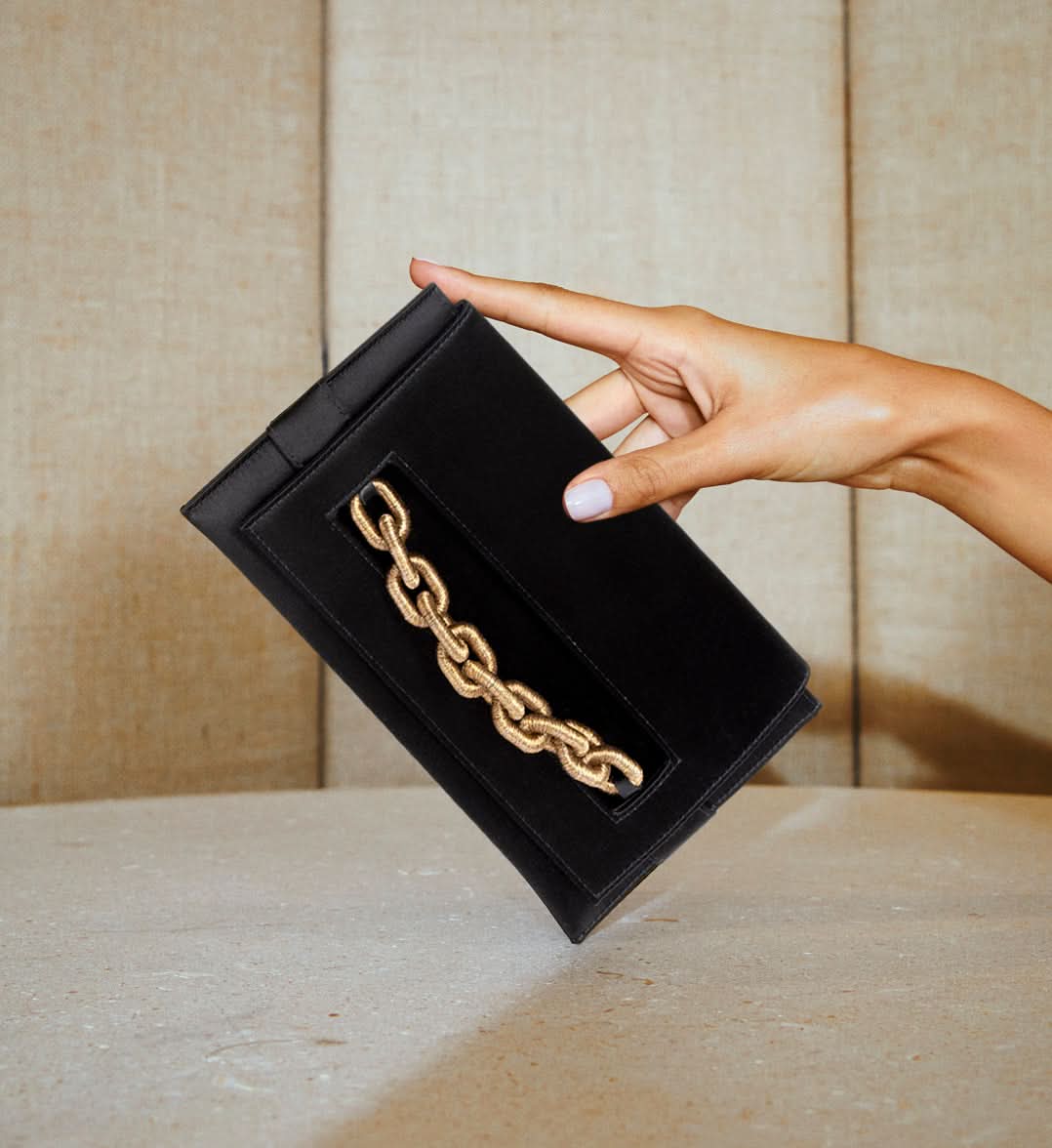 Black silk satin Catena Envelope Clutch with gold chain embroidery held against a textured backdrop.