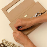 Person's hands adjusting a clasp on a beige purse with decorative chains laid beside it, showcasing style and elegance.