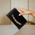 Black silk satin Catena Envelope Clutch with gold chain embroidery held against a textured backdrop.